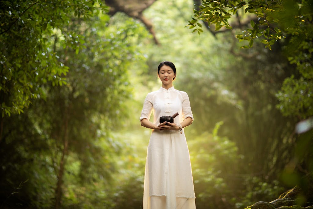 Asian woman meditating with a Tibetan singing bowl in a serene forest setting.